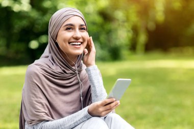 Cep telefonu için müzik uygulaması. Parkta müzik dinleyen neşeli müslüman kız, kulaklık ve akıllı telefon kullanıyor, boşluğu kopyalıyor.