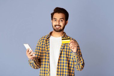 İnternetten alışveriş konsepti. Leylak arka planında cep telefonu ve kredi kartı tutan neşeli Hintli adam.