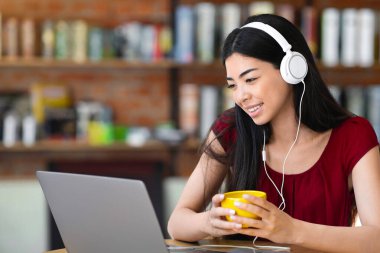 Gülümseyen Asyalı kadın laptopla ve kulaklıkla kafede oturup kahve içiyor, video konferansı yapıyor ya da internetten bilgisayar ekranına bakıyor ve gülümsüyor, E-öğrenmenin tadını çıkarıyor.