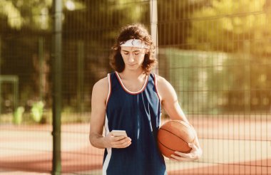 Ciddi bir genç basketbolcu oyun sonrası sahada akıllı telefon kullanıyor.