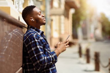 Müzik aşığı. Kapalı gözleri olan, şarkı dinleyen, duvara yaslanan rüya gibi Afrikalı bir adamın yan görüntüsü.