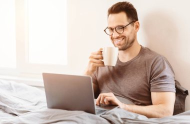 Mutlu Adam dizüstü bilgisayar kullanıyor internette geziniyor. Hafta sonları yatakta sabah kahvesi içiyor..