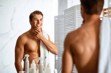 Tıraş olduktan sonra pürüzsüz yüzüne dokunan, evdeki aynanın yanında duran tıraşlı, gülen adam. Erkek Güzeli, Sabah Kişisel Bakım Programı ve Yüz Bakımı Konsepti. Omuzdan Görünüm