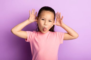 Dilini uzatan ve stüdyoda aylak aylak dolaşan şirin Asyalı çocuk portresi, pastel mor arka planda izole edilmiş.