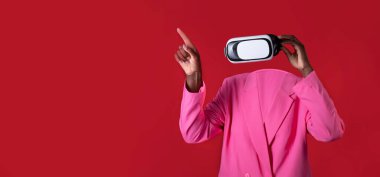 Bir kullanıcı, modern teknolojiyi ön plana çıkaran sanal gerçeklik kulaklığı takar. Kişi parlak pembe bir ceket giyer, kırmızı arka plana karşı koyu bir zıtlık yaratır, yeniliği vurgular..