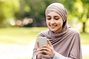 Modern akıllı telefonu kullanarak tesettürlü güzel bir Arap kızı parkta tek başına vakit geçirir, boşluğu taklit eder.