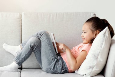 Akıllı telefon kullanan küçük Çinli kız internette oyun oynuyor. Evdeki koltukta uzanıyor. Kid ve Gadget. Panorama