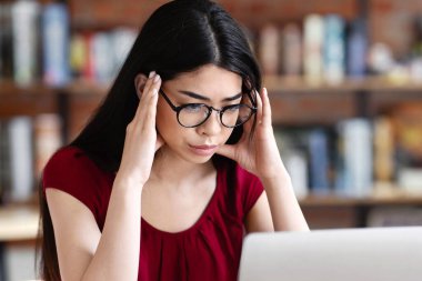 Asyalı inek öğrenci kafede dizüstü bilgisayarla çalışıyor, tapınaklara dokunuyor, konsantre olmaya çalışıyor, zor sınavlara hazırlanıyor.