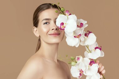 Güzel bir milenyum kadını kahverengi stüdyo arka planında orkide ile yüzüne dokunuyor. Gözünü çiçeklerle kaplayan, gülümseyerek kameraya bakan seksi genç bayan. Spa, güzellik ve sağlık konsepti