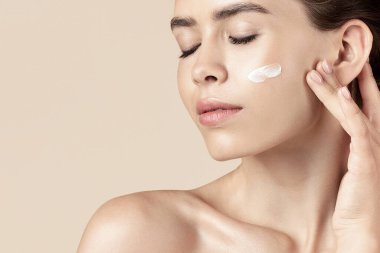 Güzellik ve sağlık konsepti. Yüzüne bej arka planda krem süren seksi genç bayan.
