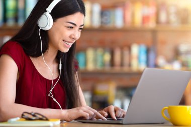Kulaklıklı gülümseyen Asyalı kız laptop ekranına bakıyor, kafede video konferans uygulaması üzerinden yabancı diller öğreniyor, boşluğu kopyalıyor.