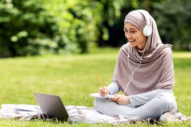Çalışkan Arap kız halka açık parkta çalışıyor, laptop ve kulaklık kullanıyor, notlar alıyor, boşlukları kopyalıyor.