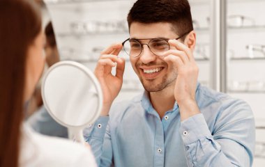 Genç bir adam bir optik mağazada aynaya bakarken gözlüğünü ayarlarken gülümsüyor. Atmosfer dostane ve görüş bakımına odaklı. Göz giyiminin önemini vurguluyor..