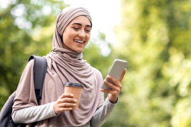 Genç Arap kadın kahve içiyor ve telefon kullanıyor, şehir parkında yürüyor, boş bir alanda.