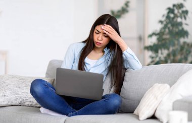 Bağımsız stres. Endişeli Asyalı kız işten sonra evde dizüstü bilgisayar başında yorgun, projeyle ilgili sorunları var, kanepede oturuyor, boş yer var.