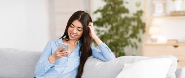 Neşeli Koreli kadın oturma odasında akıllı telefonuyla dinleniyor, hafta sonunun keyfini çıkarıyor, rahat koltukta dinleniyor, internete ya da sosyal ağlara takılıyor, arkadaşlarıyla mesajlaşıyor, mekanı kopyalıyor.