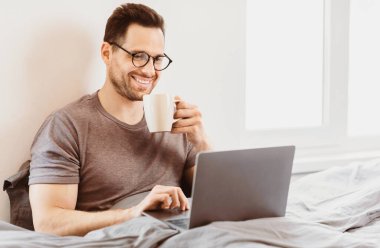 Laptopta çalışan, yatakta kahve içen neşeli, serbest çalışan adam. Evden Çalışma Konsepti.