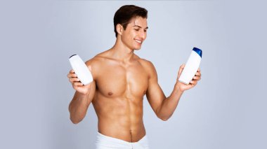 Reklam Kavramı. Gri stüdyo arka planında izole edilmiş şampuan ve duş jeli taşıyan tişörtsüz bir adam. Gülümseyen adam saç kremi ve losyon şişelerine bakıyor, kozmetik ürünleri seçiyor.