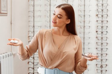 Genç bir kadın bir gözlük dükkanında duruyor, bir elinde gözlük, diğer elinde lens var. Potansiyel bir müşteriye seçeneklerini gösteriyor gibi görünüyor..
