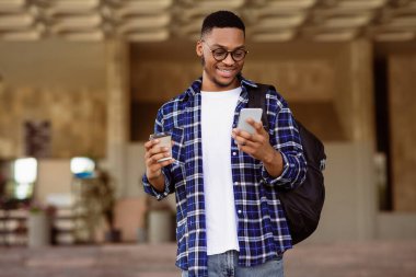 Gençlik Konsepti. Neşeli siyah hippi gözlüklü ve sırt çantalı kahve ve cep telefonuyla dışarıda duruyor.