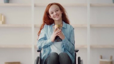 Parlak kızıl saçlı bir kız, bir tekerlekli sandalyede oturmuş göğsüne yakın bir kupa tutarken gülümsüyor ve başarısını ve mutluluğunu sergiliyor..