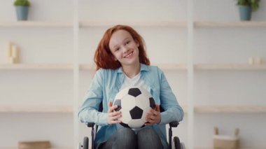 Neşeli genç bir kız tekerlekli sandalyede oturuyor ve futbol topunu neşeyle kucaklıyor. Açık mavi bir gömlek giyiyor ve uzun kızıl saçları var. Parlak kapalı alanda arka planda raflar var..