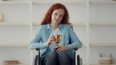 Tekerlekli sandalyede oturan kızıl saçlı bir kız gururla kupa sergiliyor. Parlak odada onun başarısını ve neşesini simgeleyen raflar yer alıyor..