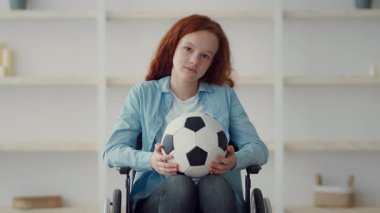 Tekerlekli sandalyede oturan genç bir kız, bir futbol topunu nazikçe kucaklıyor. Açık mavi bir gömlek giyiyor ve kıvırcık kızıl saçları var. Sıcak ve davetkar bir ortamda spora olan tutkusunu gösteriyor..