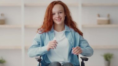 Canlı kızıl saçlı neşeli bir kız tekerlekli sandalyede oturuyor. Konuşurken sıcak bir şekilde gülümsüyor. Arka planda temiz ve modern bir iç mekan var..