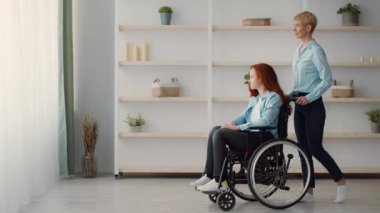 Bakıcı, tekerlekli sandalyedeki bir kızı nazikçe parlak bir pencereye doğru iter. Odada minimalist bir dekor var, doğal ışıkla dolu sıcak ve davetkar bir atmosfer yaratıyor..