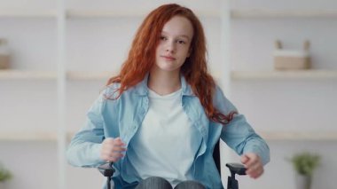 Uzun kızıl saçlı bir kız minimalist dekorlu iyi ışıklandırılmış bir odada tekerlekli sandalyede oturuyor. Etrafında dolaşırken rahatlamış görünüyor. Kendine güveni tam..