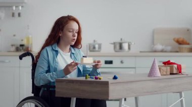 Tekerlekli sandalyede oturan kızıl saçlı genç bir kız, güzel ışıklandırılmış bir mutfakta pastasının tadını çıkarıyor. Renkli parti dekorasyonları masayı süsleyerek festival atmosferine katıyor..