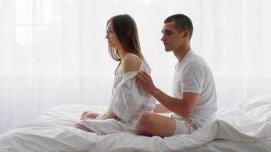 Genç bir çift, yumuşak ışıklı bir yatak odasında bir yatakta birlikte oturur. Erkek, kadının omuzlarına nazikçe masaj yapar. Huzurlu bir ortamda sevgisini ve samimiyetini gösterir..