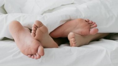 Genç bir çift yatakta dinlenir, ayakları yumuşak battaniyenin altında şakacı bir şekilde birbirine dolanır. Samimi atmosfer, huzurlu bir sabahtan zevk alırken aşklarını ve bağlantılarını yansıtıyor..