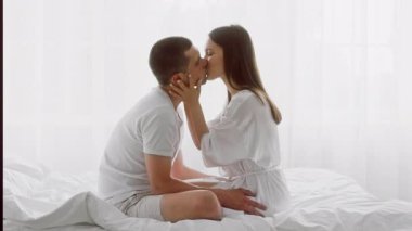 Genç bir çift, parlak bir yatak odasında yakın bir şekilde oturur, aşk ve yakınlık anlarını paylaşırlar. Gülüşleri, birbirlerinin varlığından keyif alırken paylaştıkları derin bağı yansıtıyor..