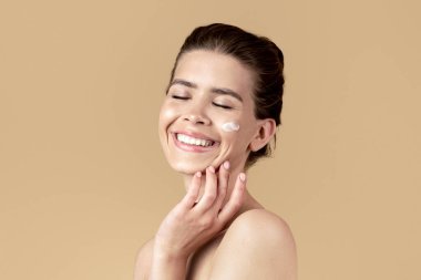 Güzellik ve deri sağlığı konsepti. Bej renkli stüdyo geçmişine yüz kremi süren mutlu genç bayan.