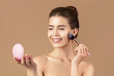Dekoratif makyaj yapmak için kozmetik fırça kullanan çekici genç bayan, kahverengi stüdyo arka planında aynaya bakıyor. Milenyum kadını yüzünde bir gülümsemeyle kızartıcı sürüyor.