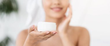 Yüz kremi reklamı. Havluya sarılıp krem kullanan mutlu Asyalı kadın. Güzel kadın ellerinde yüz kremi olan kavanoza odaklan, sabahları duştan sonra yüzüne nemlendirici sür.