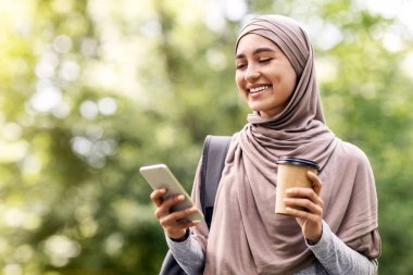 Sırt çantasıyla tesettürlü, kahve içen, telefon kullanan şehir parkında yürüyen boş bir kız.