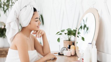 Güzel Asyalı bir kadının sabah aynanın karşısına geçip iç mekan makyajı yaptığı yan manzara. Havluya sarılı genç bir bayan yatak odasındaki aynaya bakıyor, duştan sonra sabah rutinini yapıyor.