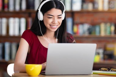 Cheeful Koreli serbest çalışan kız laptopta çalışıyor ve kafede kulaklıkla müzik dinliyor bilgisayar ekranına bakıyor ve gülümsüyor, boş bir alan arıyor.