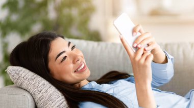 Genç Asyalı kadın evde akıllı telefon kullanıyor, rahat bir kanepede uzanırken arkadaşlarıyla mesajlaşıyor, boş bir manzarası var.