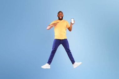 Mobil uygulama reklamı. Heyecanlı genç Afrikalı Amerikalı adam mavi stüdyo arkaplanının üzerinden atlıyor, boş ekranlı modern akıllı telefonu gösteriyor, web sitesi ya da uygulaması için model, tam boy