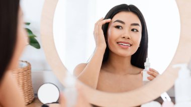 Gülümseyen Asyalı genç bir kadın yüzüne serum uyguluyor, aynaya bakıyor. Evdeki aynanın önünde sabah rutinini yapan mutlu bir kadın şakaklarına yüz serumu uyguluyor.