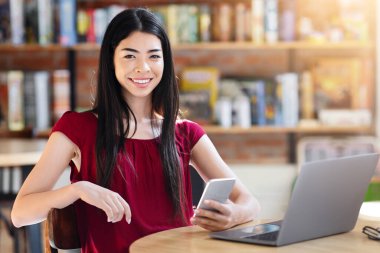 Kafede dizüstü bilgisayar ve akıllı telefonla oturan gülümseyen Koreli kız uzaktan çalışmanın keyfini çıkarıyor.