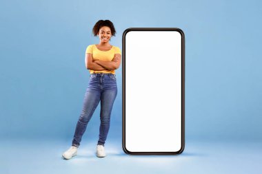 Güzel bir Afro-Amerikalı kadın, mavi arka planda beyaz ekranı boş, büyük bir akıllı telefonun yanında duruyor. Model için yer var. En yeni mobil uygulama, web sitesi, reklam kavramı