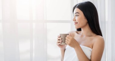 Hayalperest Asyalı genç kadın elinde bir fincan sabah kahvesi ile evdeki pencereden dışarı bakıyor, boş yer. Havluya sarılmış, duştan sonra çay içen, pencerenin yanında duran, yakın çekim yapan gülümseyen kadın.