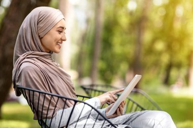 Başörtüsü takan çekici Arap kadın dijital tablet kullanıyor, parkta, kenarda oturuyor, boşluğu kopyalıyor.