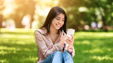 Şehir parkında akıllı telefondan müzik dinleyen mutlu Asyalı kadın çimenlikte dinlenirken şarkıyı seçiyor.