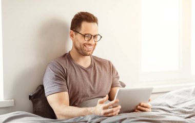 Mutlu Adam Tablet Bilgisayar kullanarak Evdeki Yatakta Oturan İnterneti tarıyor. Modern Yaşam Tarzı ve Gadget Kavramı